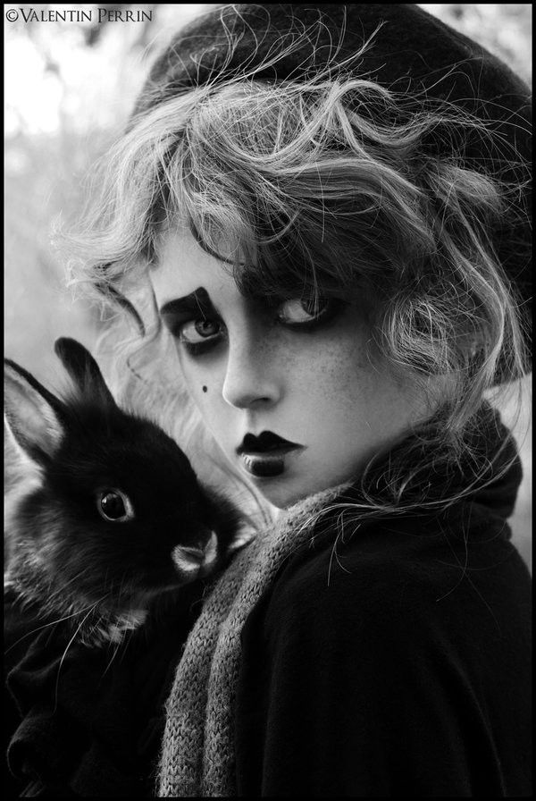 a black and white portrait of a girl with dark hair wearing a beret and a scarf. she has a dark makeup look and is holding a black rabbit close to her chest. her eyes are looking away from the camera. the photo is styled with a dramatic and artistic flair.