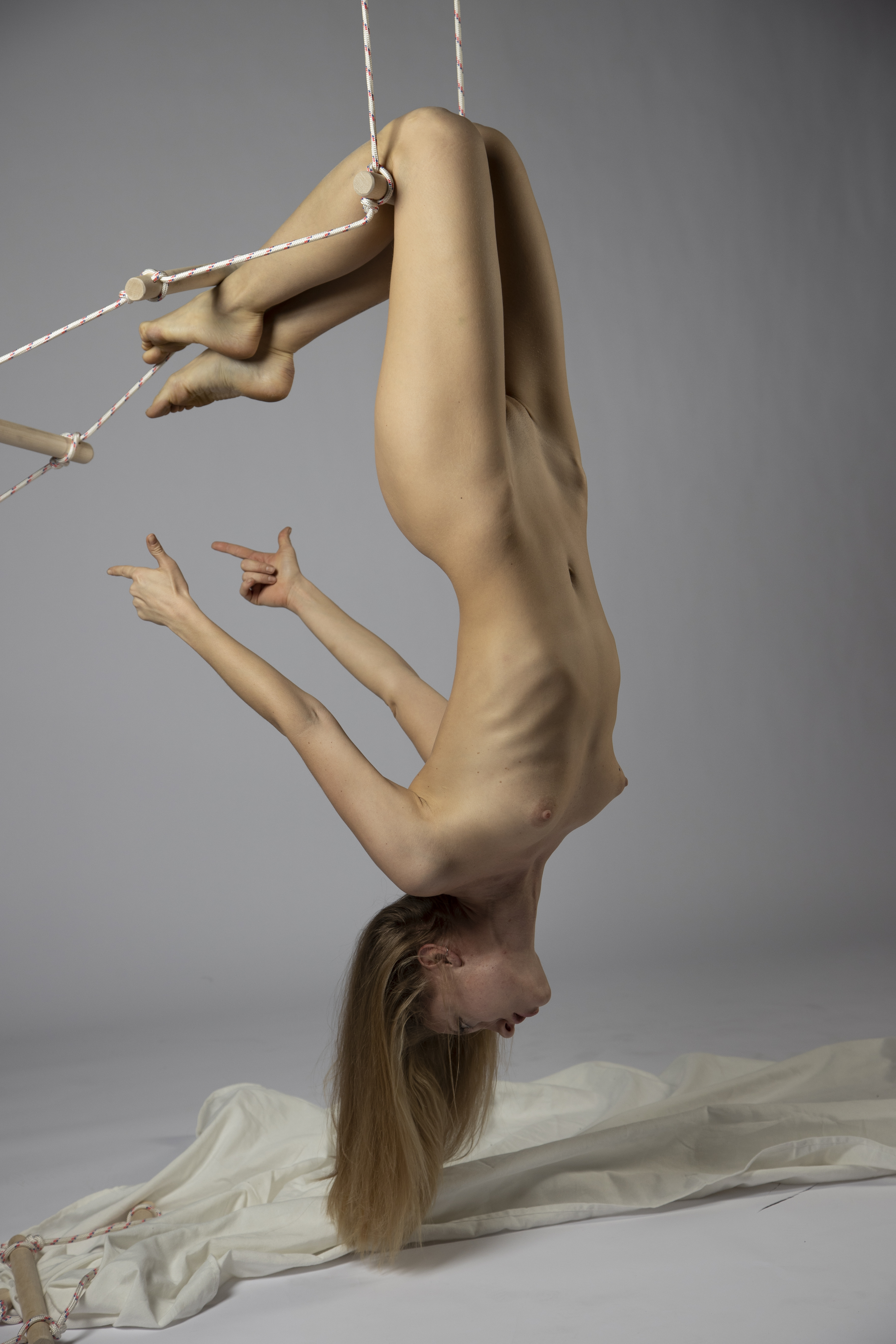 a nude woman hangs upside down from a rope ladder in a studio setting, performing an aerial acrobatic pose. she is supported by the ladder with her legs, and her arms are extended, pointing her fingers. the background is a neutral gray, and there is drapery on the floor near her head. her blond hair hangs straight down towards the drapery.
