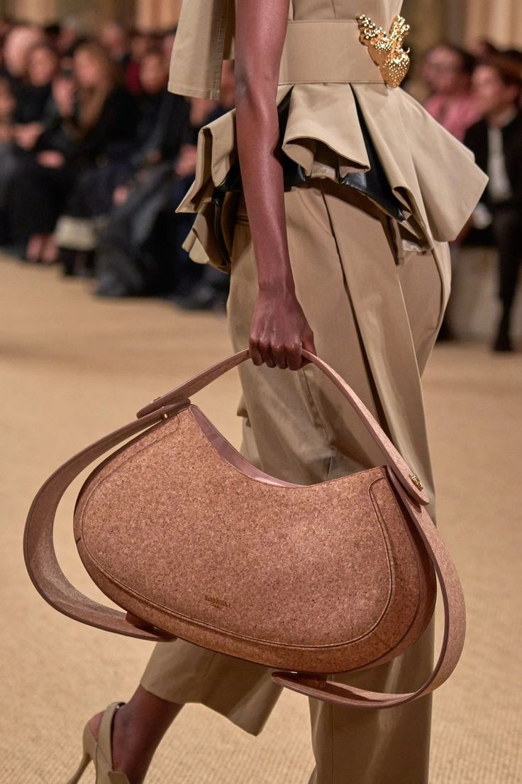 a woman is modeling a cork handbag on a runway. she is wearing a beige outfit with a matching belt with a large gold buckle. she is wearing a large cork handbag that she is holding in her hand. the handbag is a contemporary style. she looks chic and elegant in this outfit.