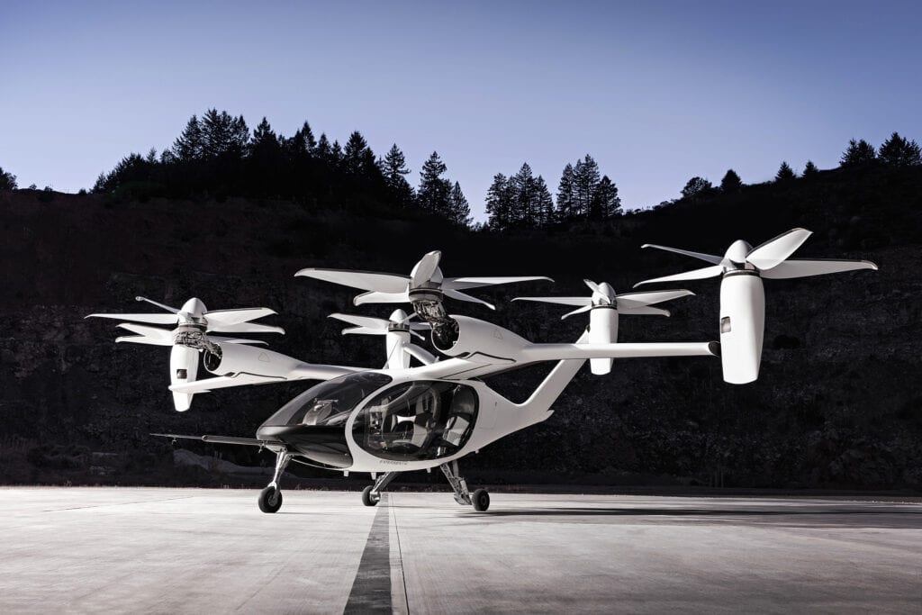 a white airbus cityairbus nextgen air taxi aircraft sits on a concrete surface in front of a dark green hill.  it is facing towards the camera with the propellers still. the sky is light blue.  the aircraft is a futuristic design that looks like a cross between a helicopter and a drone.