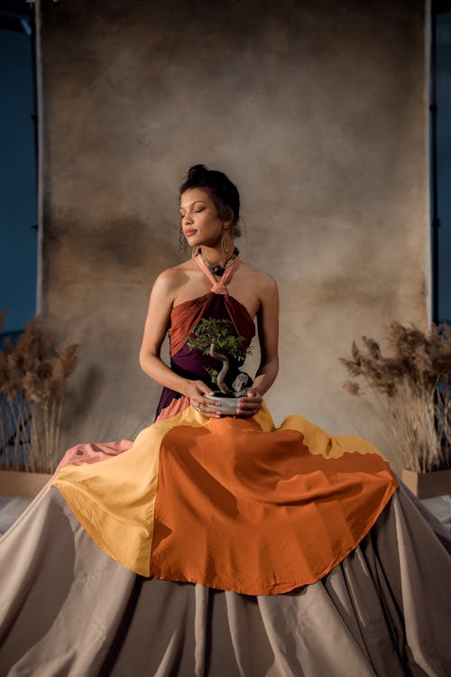 a young woman with dark hair and olive skin sits calmly on a beige fabric covered platform. she is wearing a multicolored maxi dress with shades of orange, yellow, and a deep purple bodice. the dress is a halter neck style with a flowing skirt. the model holds a small bonsai tree in a white pot. she is wearing earrings. the background is a textured neutral color, with dried plants on either side of her adding to the serene atmosphere. the overall mood is peaceful and stylish. the image is professionally lit and appears to be a fashion photograph.