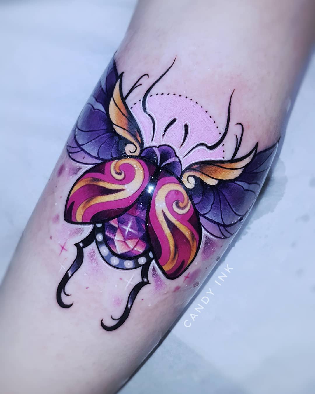 a person's forearm with a fresh neotraditional tattoo of a stylized beetle. the beetle has large, purple wings with gold accents, and a pink and gold body. a large jewel is depicted on the beetle's back. the tattoo is surrounded by small, white dots.
