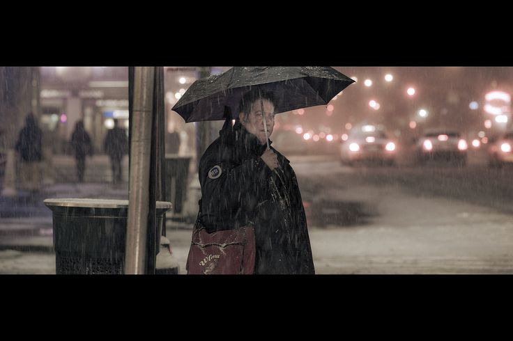 a person stands under a black umbrella in the snow on a city street at night. the image is blurry and atmospheric.  the background shows traffic lights, cars, and other people walking. the person appears to be waiting at a bus stop, or similar location. their jacket is dark, and they are carrying a dark red messenger bag. the overall mood is cold and somewhat somber.