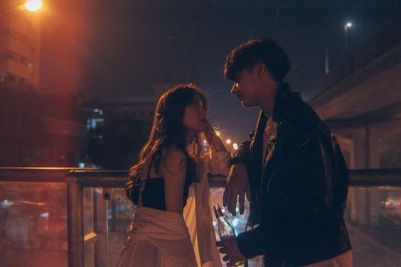 a young couple stands on a bridge at night, illuminated by the warm glow of streetlights. the city lights twinkle in the background, creating a romantic atmosphere. the man, wearing a black leather jacket, leans against the railing while looking at the woman, who is dressed in a white shirt and black top.  she gazes back at him, her hand resting on her chin. the scene conveys a sense of intimacy and connection.