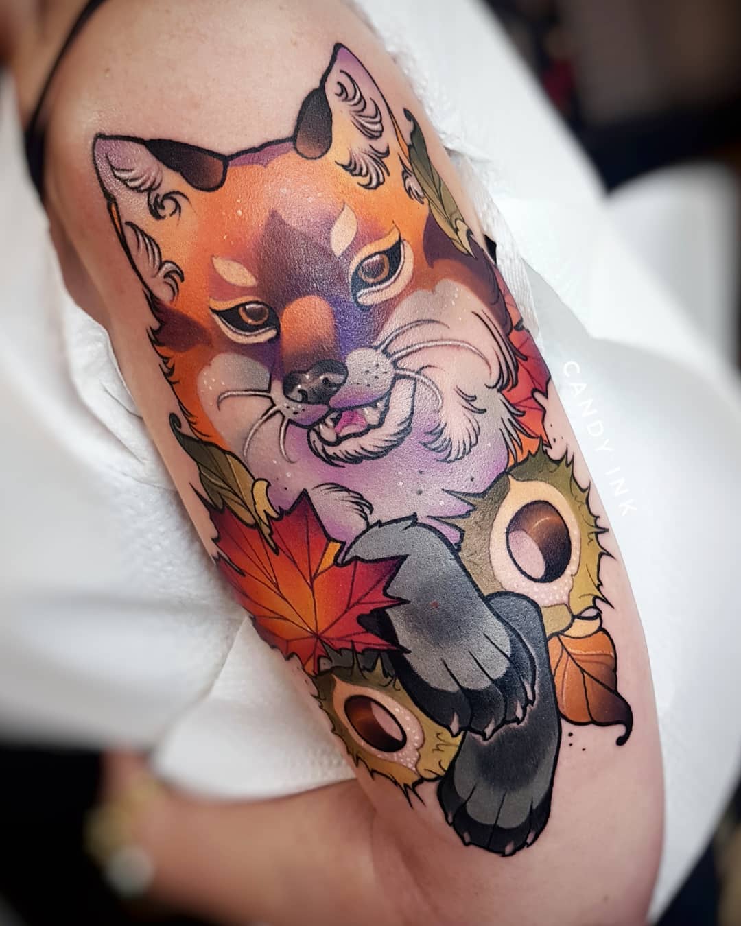 a closeup photograph showcasing a vibrant neotraditional tattoo of a fox on a woman's upper arm. the fox is depicted in a playful pose, surrounded by autumnal elements such as fall leaves and acorns. the tattoo is rendered with bold outlines and a warm color palette, typical of the neotraditional tattoo style.