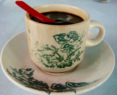 a closeup shot of a coffee cup on a saucer. the cup is white with green floral designs, and the saucer has similar designs. the cup contains dark coffee, and a red spoon is resting on the rim.  the background is blurred.