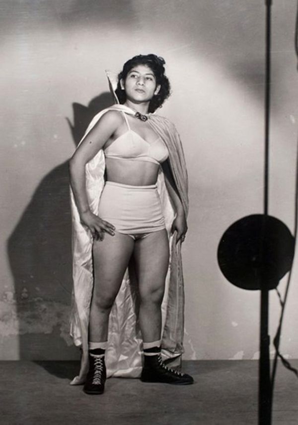 a black and white studio portrait of a female wrestler, possibly from the 1950s. she is wearing a wrestling costume consisting of a lightcolored twopiece outfit,  hightop boots, and a long cape or robe draped over her shoulders. she has dark hair and a serious expression, standing confidently with her hands on her hips. the background is a plain studio wall with a shadow cast to the left and a reflector to the right, typical of a professional photographic setup. the image conveys a sense of strength and determination.