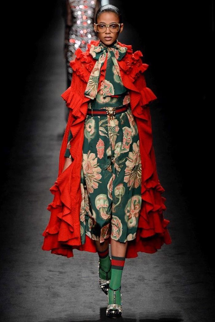 fashion model walks the runway in a striking gucci outfit. she wears a long, ruffled red cape over a dark green dress with a floral pattern. the dress has a cinched waist with a gold buckle belt. she accessorizes with green and red striped socks and embellished black shoes. the model also wears glasses. the background is dark and blurry, focusing attention on the model and her outfit. the overall impression is one of high fashion and bold style.