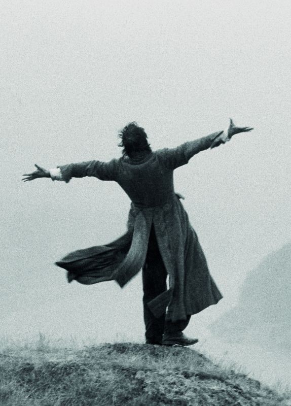 a man in a long coat stands on a hilltop with his arms outstretched, as if embracing the wind. the image is in black and white, giving it a dramatic and moody atmosphere. the sky is overcast, and the wind is blowing the man's coat behind him. the overall feeling of the image is one of solitude and freedom.