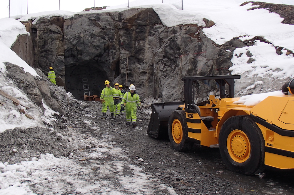 workers in hardhats walk out of a tunnel, a yellow excavator stands in the foreground, with snow covered rock faces on either side.  the workers are likely construction workers or miners.