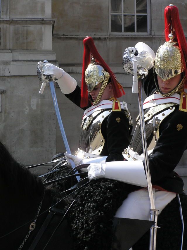 two soldiers from the household cavalry are mounted on horses, wearing their distinctive ceremonial uniforms, including silver helmets with red plumes. each holds a drawn sword. they are positioned close together, filling much of the frame.