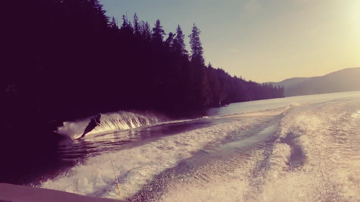 the image shows a person waterskiing on a lake at sunset.  the skier is in the air performing a jump, creating a large spray of water. the wake of the boat is prominently featured in the foreground, leading the eye towards the skier and the shoreline in the background. the background consists of a dense forest line along the lakeshore, and hills or mountains in the distance, bathed in the warm light of the setting sun. the overall feel of the image is one of summer activity and outdoor recreation. the viewpoint appears to be from a boat following the skier.