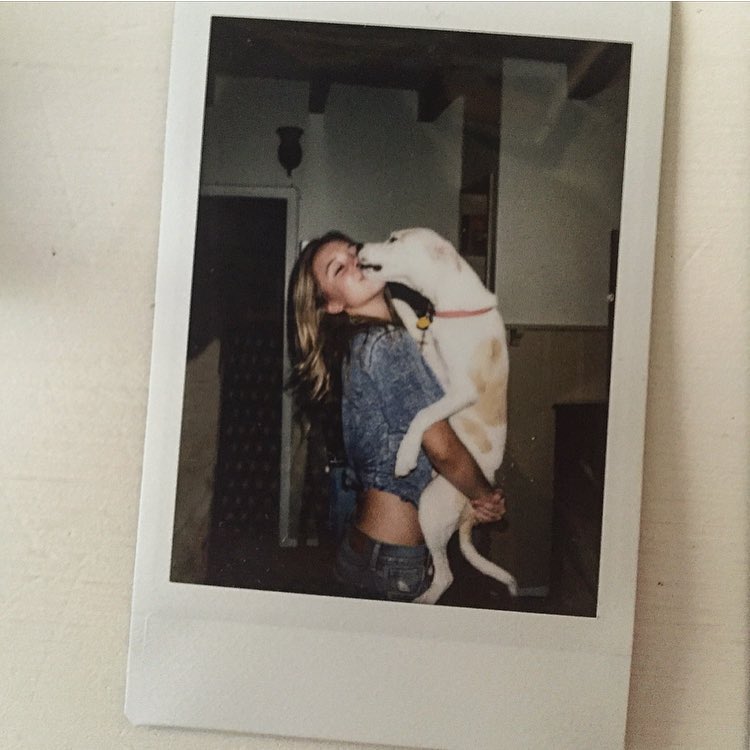 a polaroid photograph shows a young woman with long brown hair hugging and kissing a white dog. the woman is wearing a denim shirt and jeans. they appear happy and affectionate. the setting is an indoor space that looks like a home, and the lighting is dim. the overall feel is casual, candid, and nostalgic. the photo has a vintage, polaroid aesthetic.
