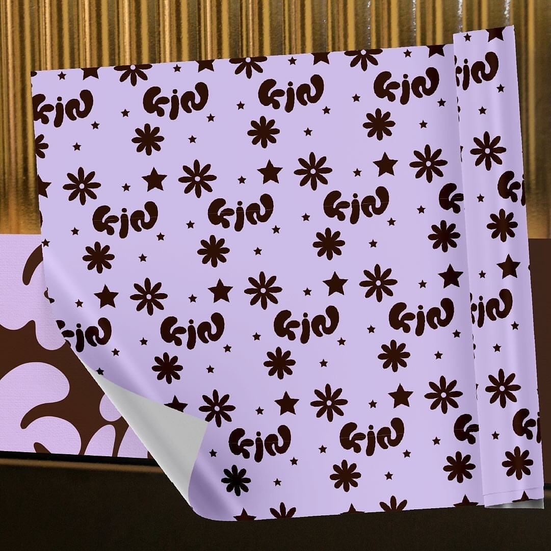 the image shows a sheet of lilac wrapping paper with a brown flower and star pattern. the left bottom corner of the paper is folded, revealing a brown surface underneath. the background features vertical golden stripes.