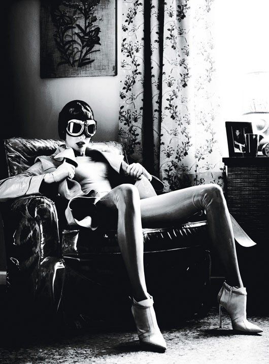 a black and white photo of a woman wearing stylish black and white clothing and a pair of aviator goggles. she's sitting on a leather chair and staring directly at the camera. she has a bold and confident expression.