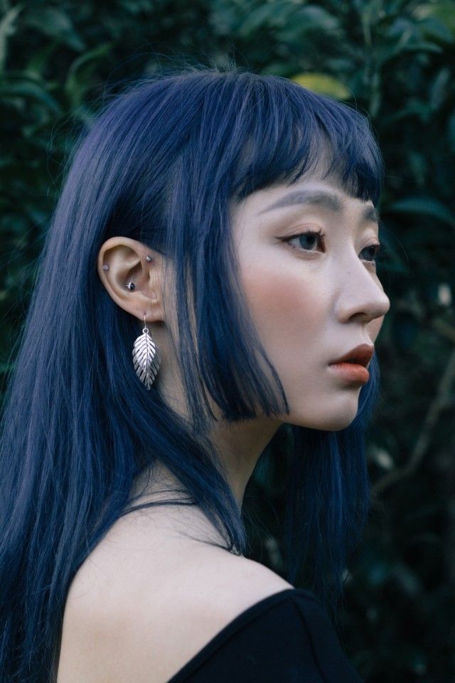 a young woman with long, dark blue hair and bangs is looking off to the side. she is wearing leaf earrings and a black shirt with a silver choker necklace. her skin is pale, and she has a dark eye shadow look with white eyeliner. her background is blurred, but green foliage can be seen.