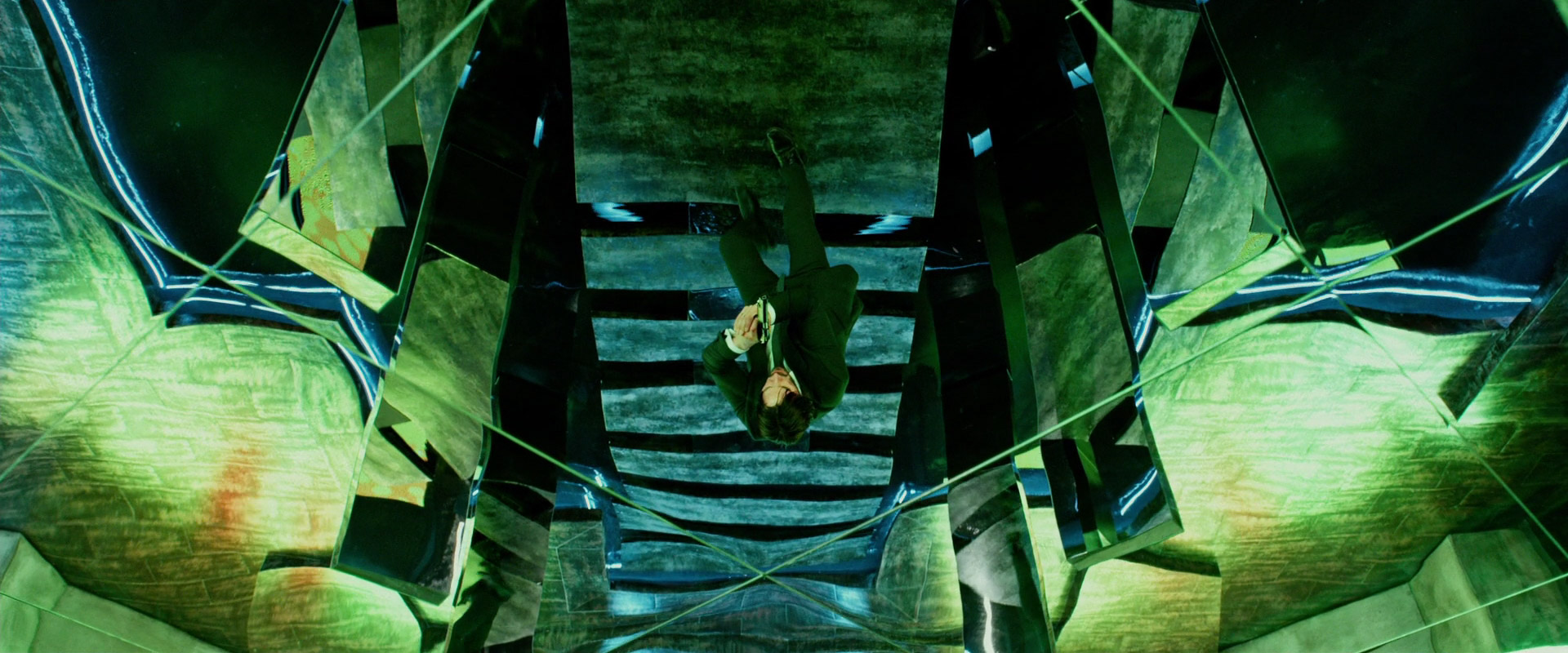 a man in a suit stands in a room with mirrored walls and ceilings, looking down at the reflection of himself below. the room is a maze of green and blue geometric shapes.