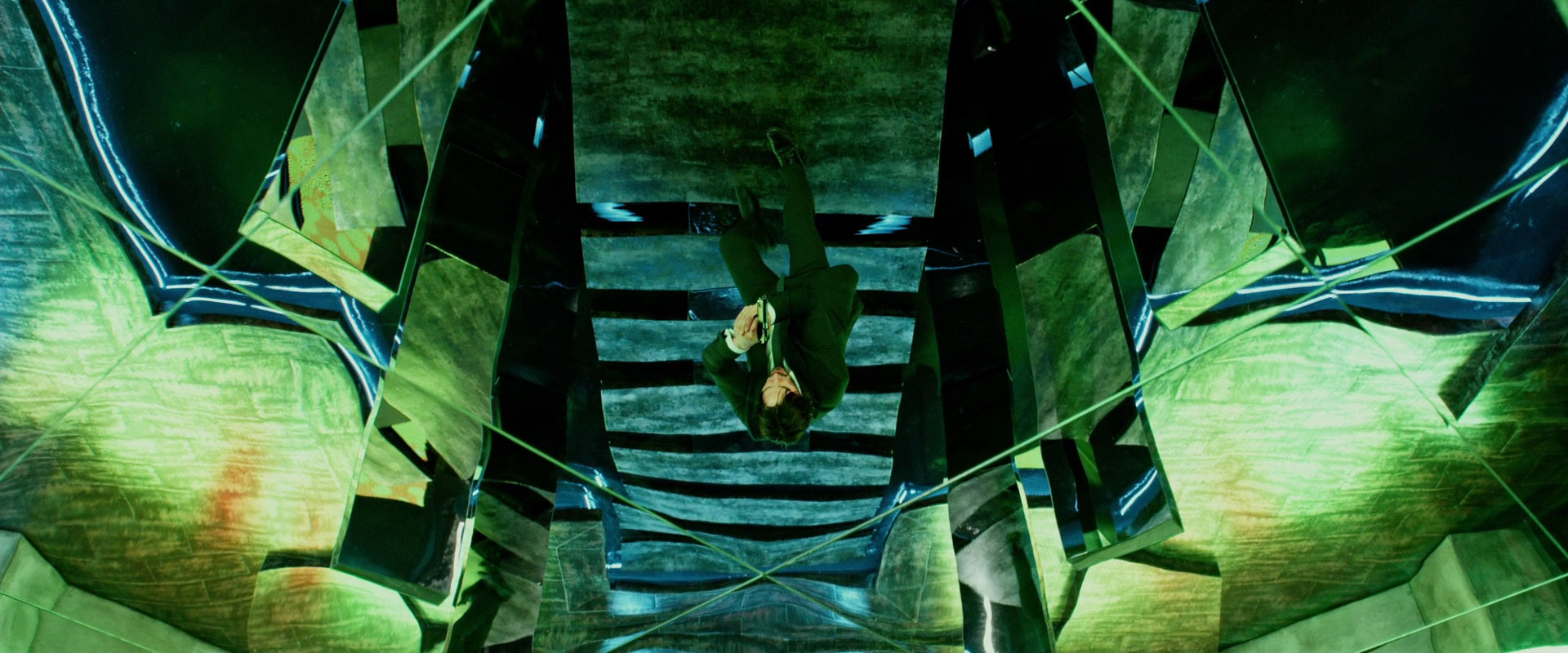 a man in a suit is reflected in a multifaceted mirrored ceiling. the mirrors create a kaleidoscopic effect, with the man appearing multiple times from different angles. the overall color scheme is green, with hints of blue and white. the lighting is artificial and bright, likely neon. the perspective is from directly below, creating a sense of vertigo.