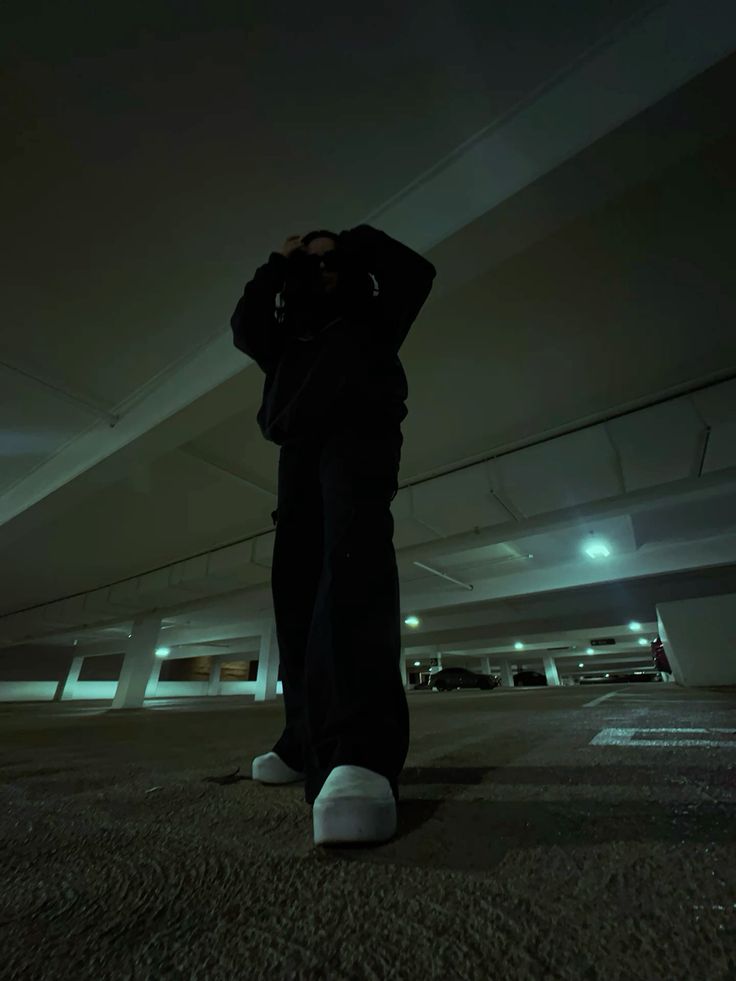 a man in a black jacket and pants stands in a dark parking garage, his silhouette illuminated by the dim lights overhead. he is looking down at his camera and holding it in his hands. he is wearing white shoes and his pants are cuffed at the bottom. he has a cool and stylish look. the background is mostly dark, with only the faint outlines of cars and other objects visible. the image has a moody, urban feel.
