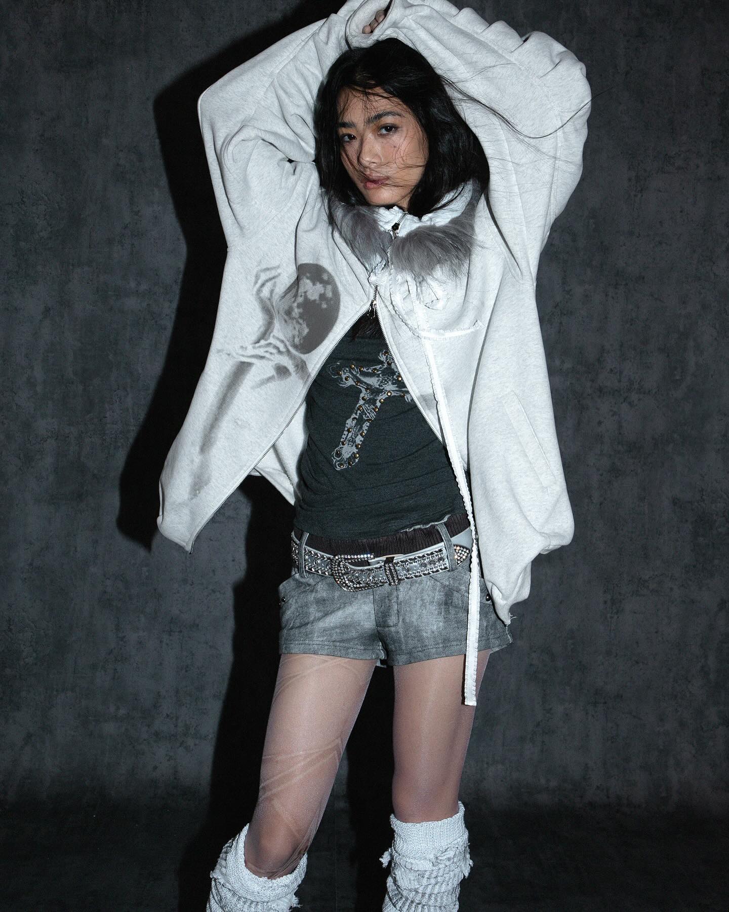 young asian woman is captured in a studio shot, her pose exuding a cool and edgy vibe. she is wearing a light gray oversized hoodie, a dark gray graphic tshirt, distressed denim shorts, sheer tights adorned with subtle markings, and white knit leg warmers.  a dark brown belt with a prominent buckle cinches her waist. her dark hair is partially pulled back, and she has a somewhat serious expression. the background is a dark gray textured wall, which contrasts with her attire. the overall mood is stylish and slightly mysterious.