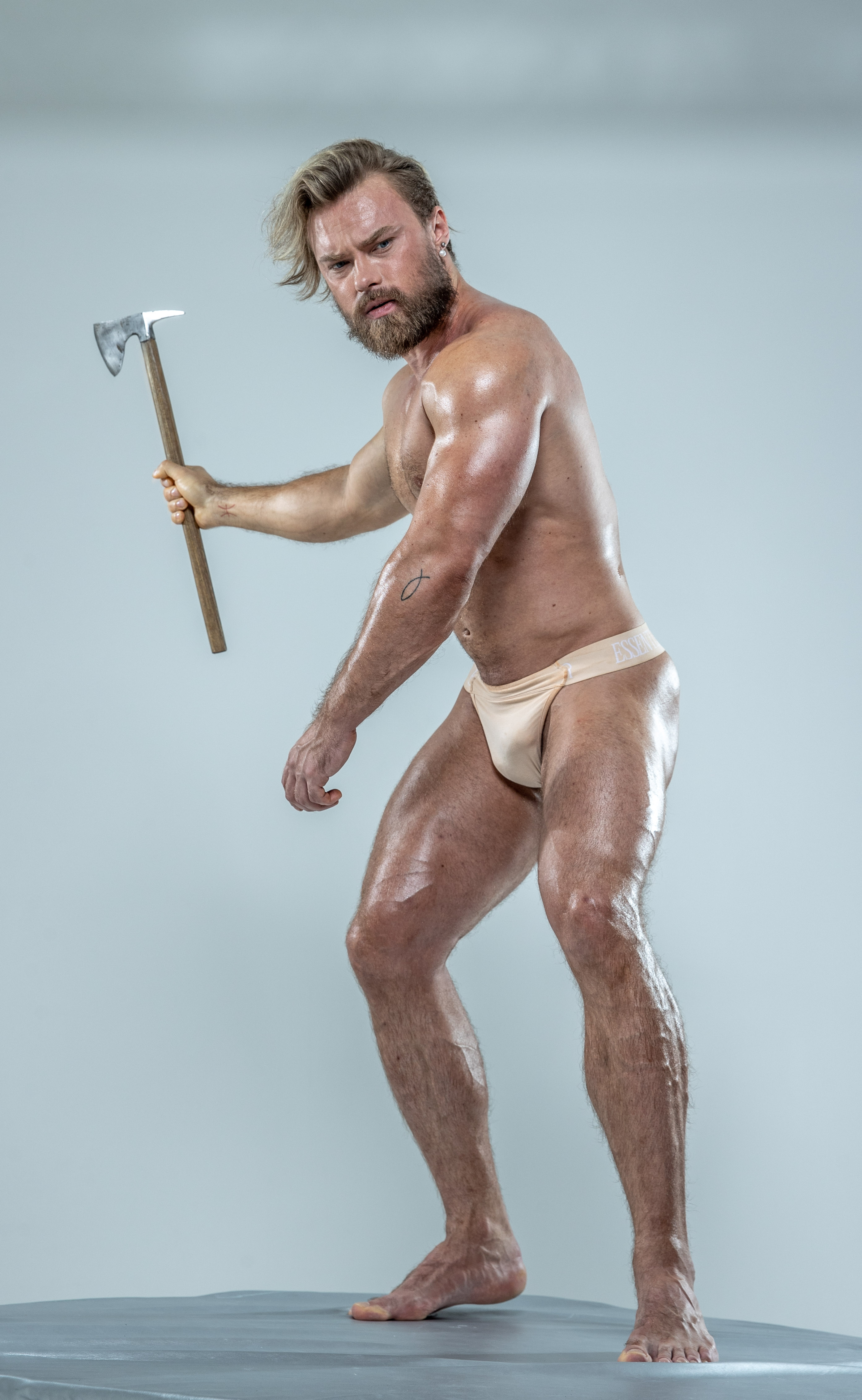 a muscular, blonde man with a beard stands on a gray platform against a light gray background. he is shirtless and wearing only a lightcolored loincloth. he holds a small axe in his right hand, raised in a defensive posture.  he is oiled and appears powerful and intimidating. his physique and the axe suggest a warrior or a figure from mythology.