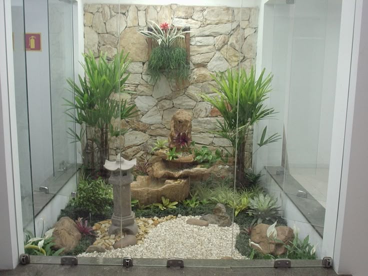 an indoor garden is enclosed by glass panels, creating a miniecosystem. the garden features a stone wall backdrop, a small waterfall feature, a lantern statue, and various plants and flowers. white pebbles cover part of the ground. a fire extinguisher sign is visible on a white wall outside the enclosure.