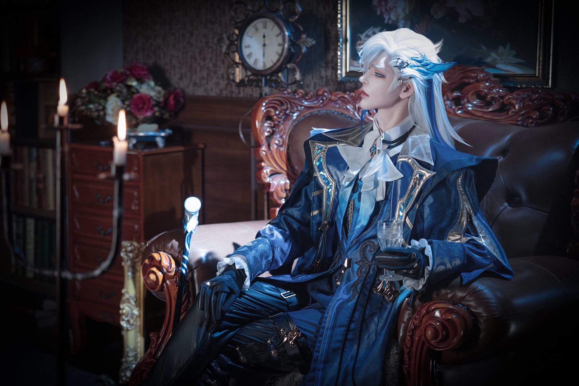 a male anime character is cosplaying in a dark and ornate room. he is sitting on a leather couch, wearing a blue jacket with gold trim and a white shirt, holding a glass. he has white hair, and is looking to the left. there are candles in the background and a bookshelf.