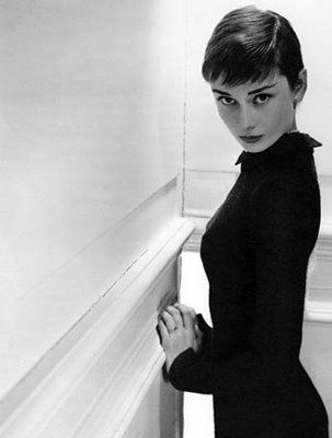 black and white photo of audrey hepburn. she is wearing a black turtleneck dress and is looking at the camera. she is standing in a doorway and is holding onto the door frame with her left hand. the background is white.