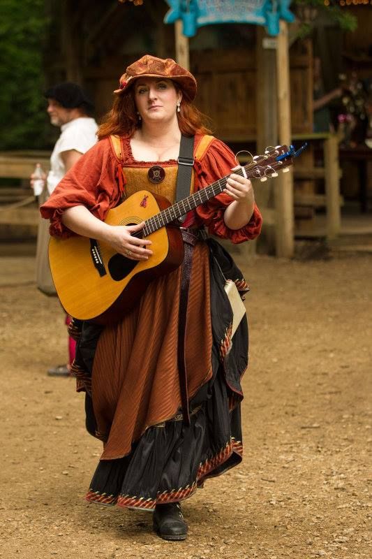 a woman dressed in a renaissance or medievalstyle costume plays an acoustic guitar at an outdoor event, possibly a renaissance fair or festival. she wears a brown hat, a puffysleeved orange top, a long brown and black skirt with red trim, and black boots. she is holding the guitar with her left hand on the fretboard and her right hand strumming the strings. the background shows a rustic wooden structure and blurred figures, suggesting a lively fair atmosphere.
