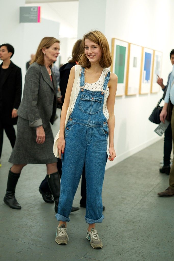 a young blonde woman smiles confidently, standing in an art gallery. she's wearing a casual yet stylish outfit: denim overalls with the legs cuffed, a white lace top, and light grey sneakers. her hair is short and styled neatly.  she's the clear focus of the image, while other people in the background are blurred, suggesting a candid street style shot.  the setting of an art gallery adds context to the scene.