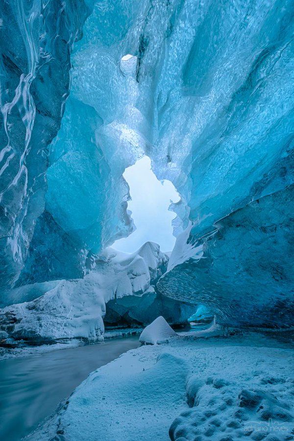 this is a breathtaking view inside a stunning ice cave. the walls of the cave are made of thick, clear blue ice that forms intriguing patterns and shapes. snow covers the floor of the cave, and a stream of water flows through it. the exit of the cave reveals a bright, snowy scene, creating a stunning contrast of light and shadow.