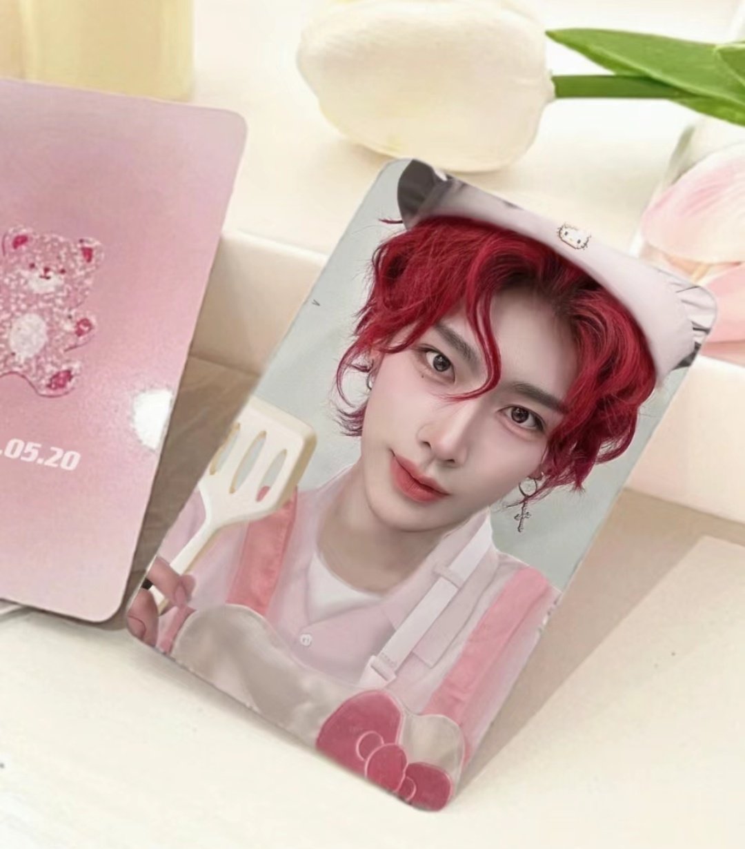 a photocard of a young asian man with bright red hair.  he is wearing a pink and white outfit.  the photocard is sitting on a pink and white surface.  there are some pink tulips in the background.