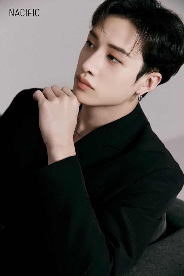 the image is a closeup portrait of bang chan, a member of the south korean kpop group stray kids. he is shown from the chest up, angled slightly away from the camera.  he's wearing a black suit jacket and has a thoughtful, almost pensive expression. his hand is gently resting on his chin. the background is plain and dark, drawing focus to bang chan. the image has a sophisticated and stylish feel, and the "nacific" logo suggests it's promotional material for the beauty brand.