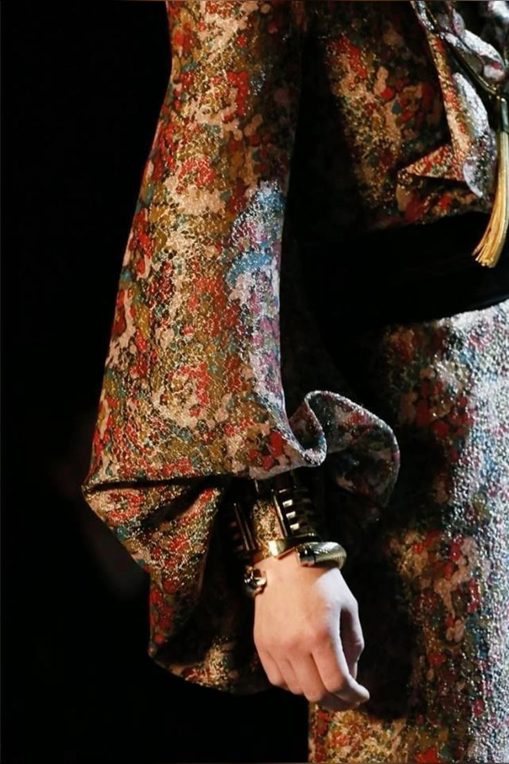 a closeup shot of a woman's arm wearing a gold bracelet and a floral patterned dress. the dress has a long sleeve with a large ruffle at the elbow. the woman's hand is visible, showing off the intricate details of the bracelet. the background is blurry and dark, drawing attention to the dress and its intricate details.