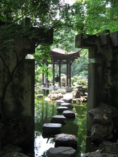 the image shows a serene scene from a chinese garden.  framed by a mosscovered stone archway, we see a traditional pavilion situated across a pond. a path of stepping stones leads towards the pavilion, inviting the viewer to cross. lush green trees and foliage surround the pond, creating a sense of tranquility. two individuals can be seen standing within the pavilion, enjoying the peaceful surroundings. the dappled sunlight filtering through the trees illuminates the scene, highlighting the vibrant green hues.