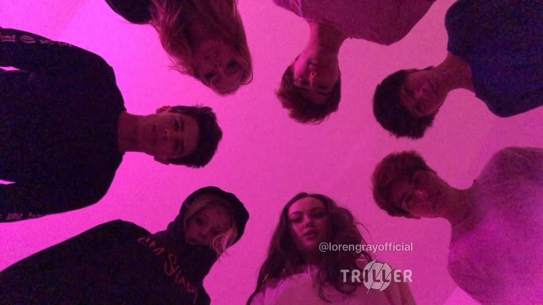 a group of six young adults are gathered in a circle, looking down at the camera in a lowangle shot with pink lighting. the triller logo and loren gray's official account handle are visible in the frame, suggesting this may be content for social media. the photo conveys a sense of friendship and unity among the group.