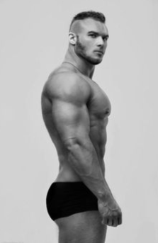 black and white image of a muscular man in underwear, posing in profile against a plain background. his physique is emphasized by the studio lighting.