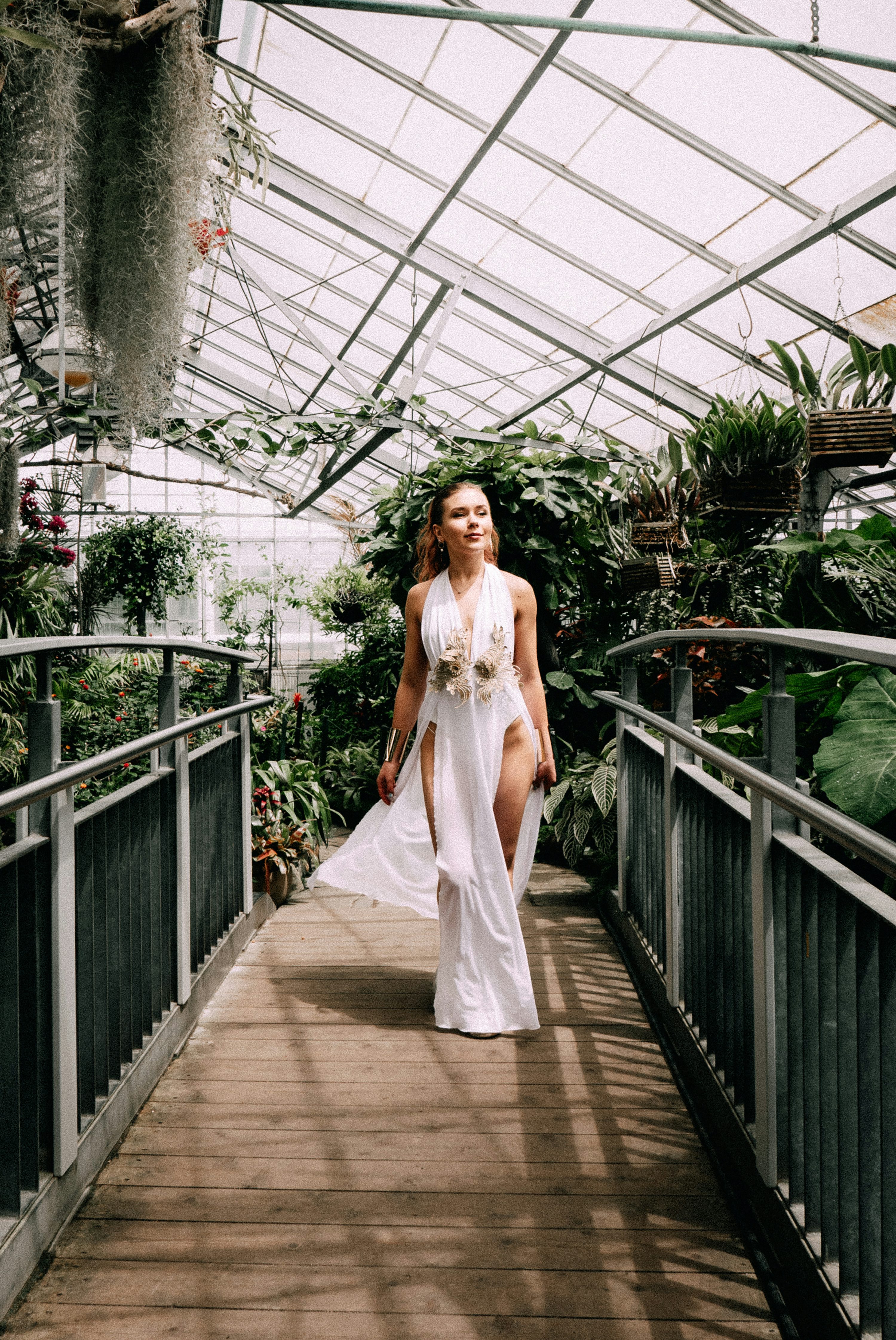 a woman wearing a white dress is walking on a wooden bridge inside a greenhouse. the greenhouse is filled with plants and has a metal frame. the bridge has metal railings on both sides. the woman is wearing a long white dress with a high slit. she is also wearing jewelry on her arm.