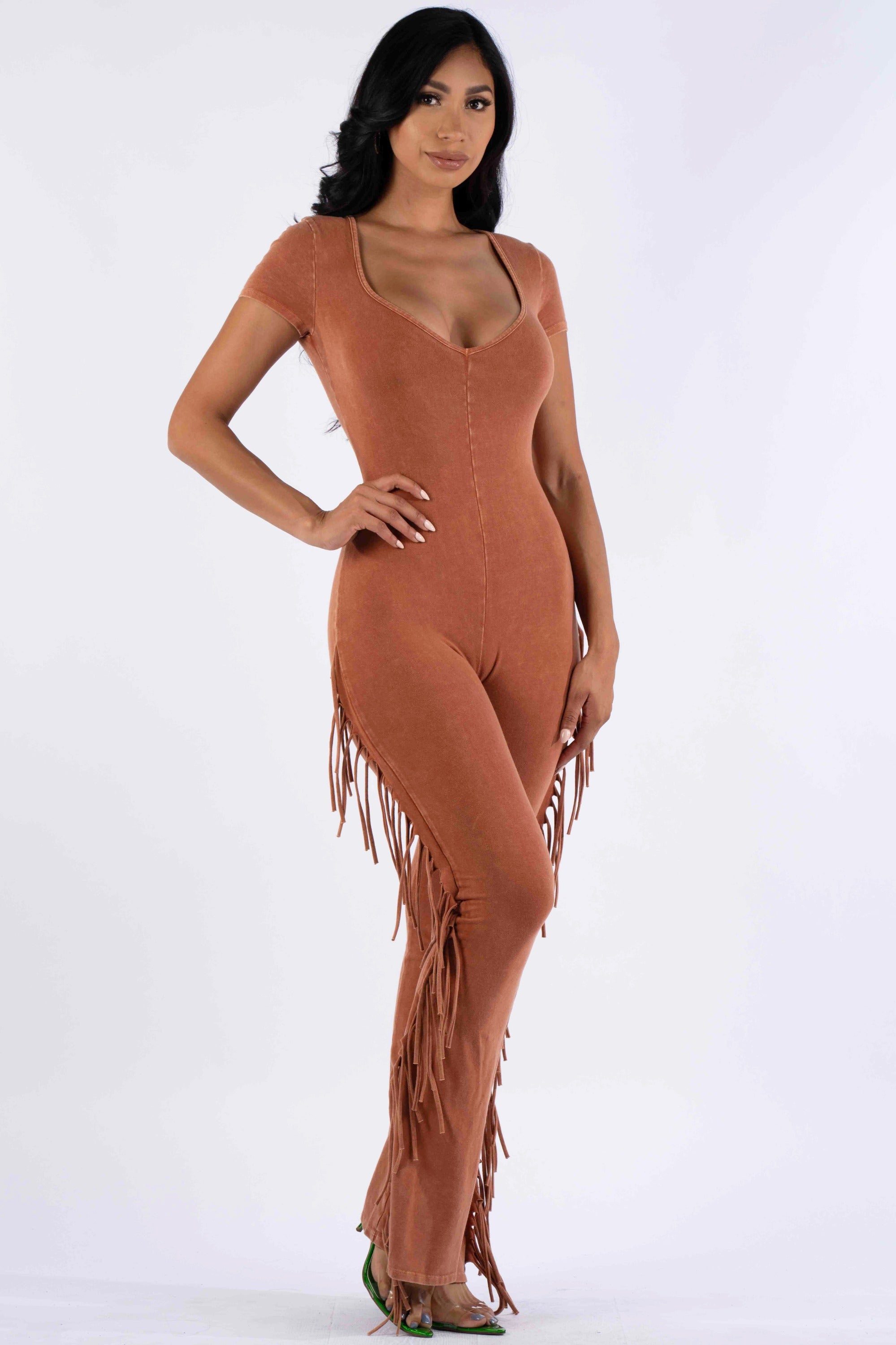 fulllength studio shot of a model wearing a terracottacolored jumpsuit. the jumpsuit has short sleeves, a vneckline, and is fitted to the body. the pants have fringe details at the sides. the model has long, dark hair and is wearing minimal makeup. she is standing against a white background and looks directly at the camera.
