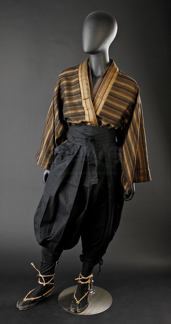 a mannequin is wearing a traditional japanese  samurai outfit.  the samurai outfit includes a dark brown and gold kimono with a black belt, black pants and sandals. the mannequin is standing with its arms at its side.