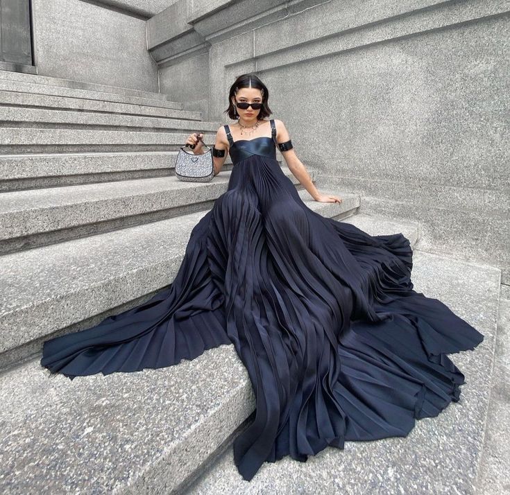 a woman is sitting on a stone staircase wearing a black, floorlength, pleated dress. she is also wearing sunglasses, a necklace, and holding a purse.