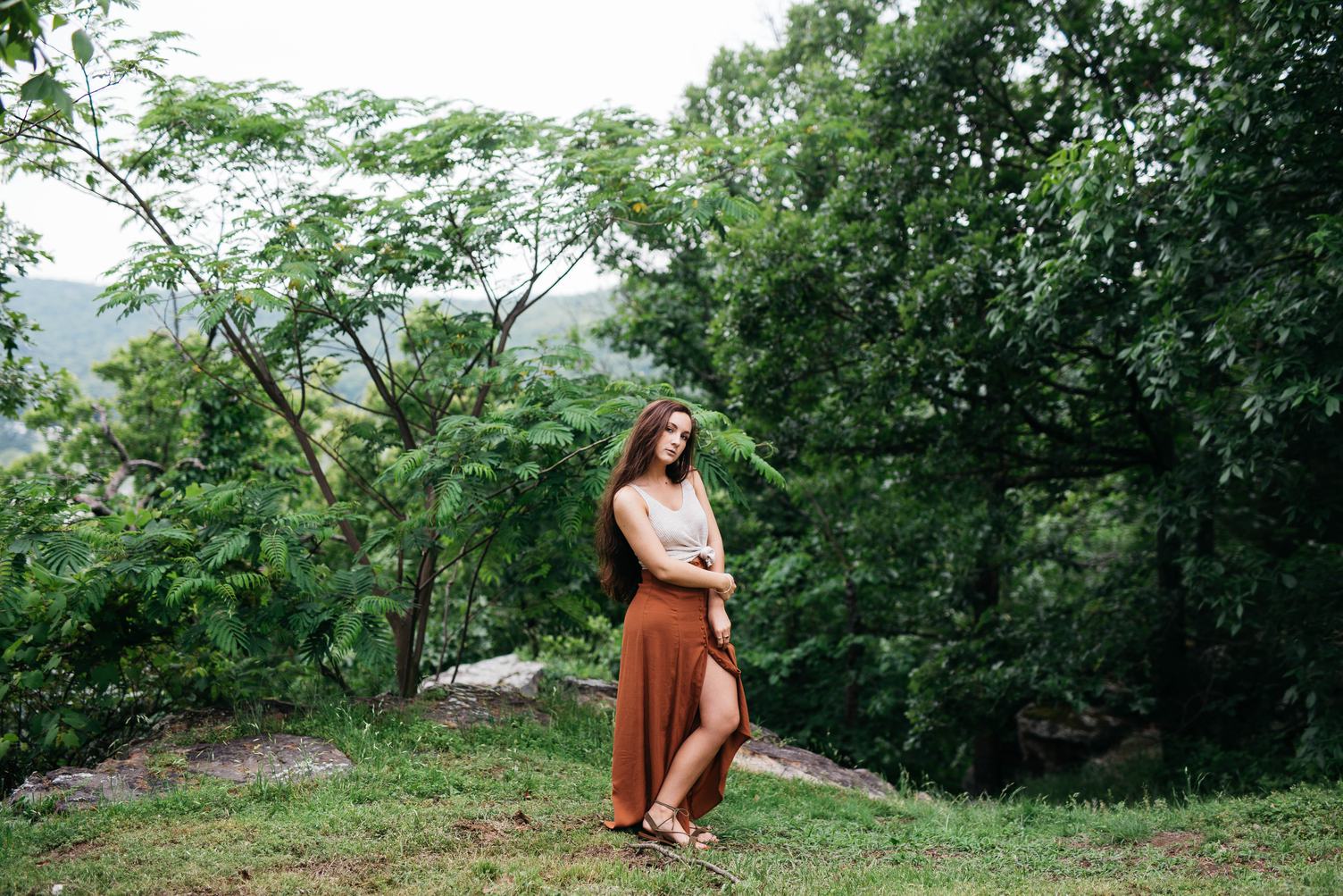 a full shot captures a woman standing outdoors, surrounded by lush greenery. she is dressed in a tan top and a long, flowing brown skirt with a slit. the woman's long hair cascades down her shoulders, and she is wearing sandals. she is posed in a natural setting, with trees and grass in the background. the overall ambiance is bohemian and serene.
