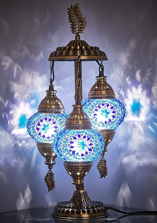 a lit, decorative turkish mosaic table lamp with three blue glass shades is displayed against a softly lit background, creating patterns on the wall. the lamp features an ornate brass stand and hanging leafshaped pendants.
