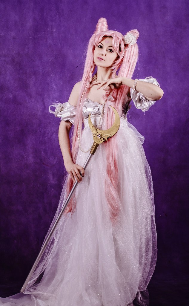 a full body portrait of a woman cosplaying as princess serenity from sailor moon. she has pink hair styled into the character's signature buns and long pigtails. she is wearing a long, flowing white dress with silver details on the bodice and sleeves. she is holding a staff with a golden crescent moon on top. she is also wearing a tiara, pearl necklace, and bracelet. the background is a purple backdrop.