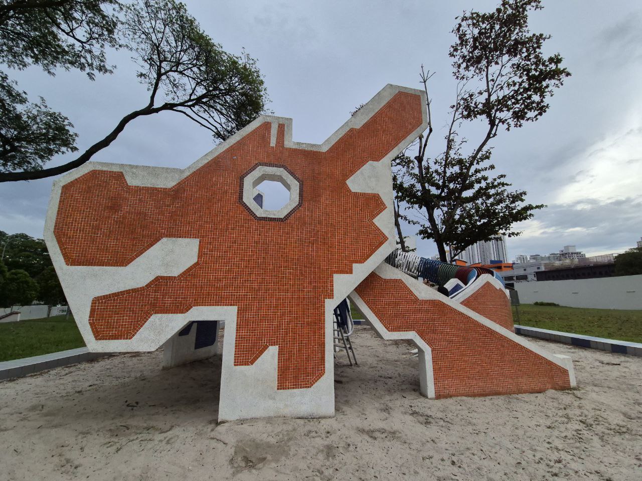 a large orange and white mosaic slide in a playground, designed to resemble a stylized animal. the slide is made of concrete and is decorated with small tiles in a brick pattern. the surrounding area is covered in sand, and there are trees in the background.