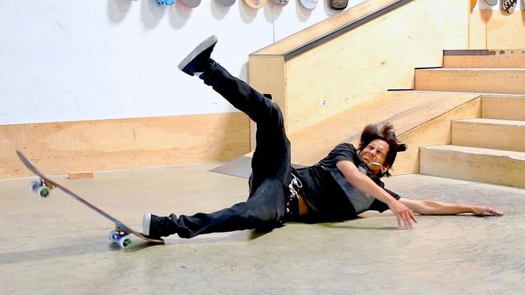 a skateboarder falls off his skateboard and lands on the ground. he is wearing black pants, a black tshirt, and a white hat. he is in a skatepark with a ramp in the background. the skateboard is still rolling on the ground.