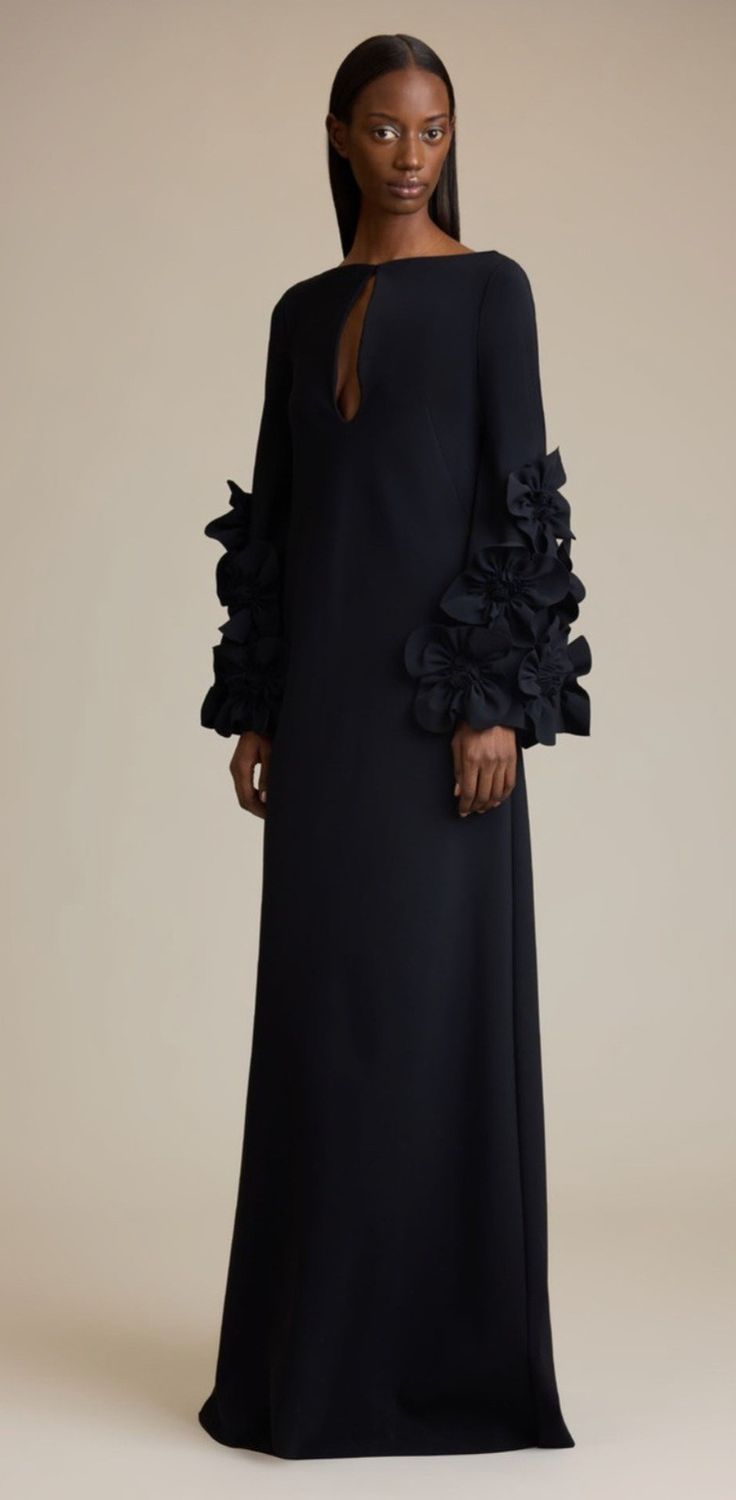 a fulllength studio shot of a darkskinned woman modeling a long, black dress. the dress has a high neckline, long sleeves with ruffled details at the cuffs, and a flowing skirt. the dress appears to be made of a smooth, possibly crepe material. the model's pose is simple and elegant.  the background is a neutral beige. the overall aesthetic is one of understated sophistication and elegance.