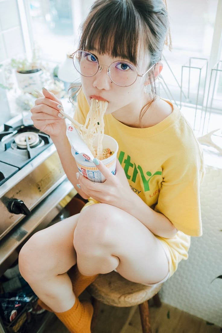 a young woman wearing eyeglasses and a yellow tshirt is sitting on a stool in a kitchen, eating noodles from a cup with chopsticks. she has her bangs down and is looking at the camera. a gas stove is visible to her left.