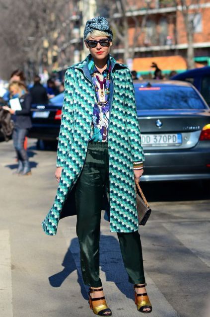 a stylish woman walks down a city street. she is wearing a teal geometric patterned coat, dark green satin pants, a patterned shirt, a turban, sunglasses, and gold heeled sandals.  she carries a brown handbag. the background shows other pedestrians and city buildings. the overall impression is one of sophisticated and trendy street style.