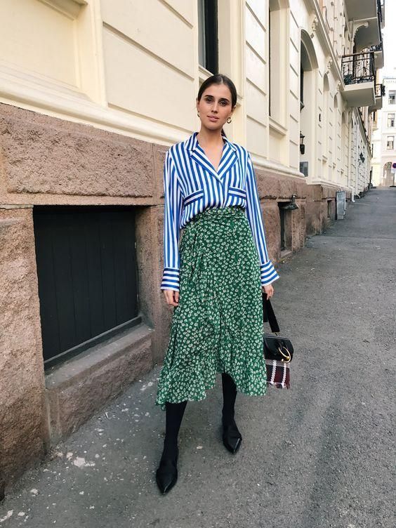the image showcases a stylish woman posing on a city street. she's wearing a blue and white striped shirt layered over a green floral wrap midi skirt.  her accessories include black tights, pointedtoe heeled shoes, and a small crossbody bag with a plaid detail.  the architectural background suggests an urban setting, and her overall style is elegant and contemporary. the lighting and setting indicate it may be autumn.